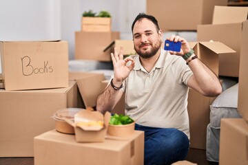 Wall Mural - Plus size hispanic man with beard moving to a new home holding credit card doing ok sign with fingers, smiling friendly gesturing excellent symbol