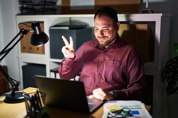 Wall Mural - Plus size hispanic man with beard working at the office at night showing and pointing up with fingers number two while smiling confident and happy.