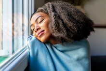 Young Woman Wrapped In Blanket Leaning On Window At Home