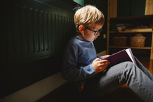 Boy Wearing Eyeglasses Reading Book Sitting On Floor At Home