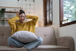 © wichayada - Hand on temples of young unhappy sadness Asian girl sitting on sofa. She is feeling not very good due to her sickness and having a headache. Seen from the front. Stress and sickness concept.