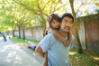 Smiling Asian man walking along alley in park with little girl. Happy father in casual clothes resting carrying daughter on his back and looking straight. Fatherhood and spending time together concept