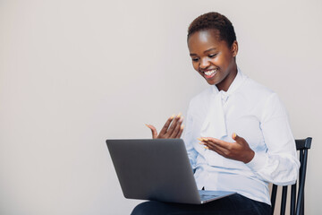 Portrait of attractive cheerful satisfied african woman sitting in chair watching webinar, working online on laptop, talking in video chat isolated over white