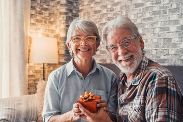 Wall Mural - Portrait of two cute and old seniors at home having fun together. Mature man  giving a gift at his wife for Christmas or anniversary. Surprised pensioner woman looking at the camera..