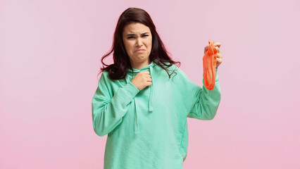 Wall Mural - young and disgusted woman in turquoise hoodie holding stinky socks isolated on pink