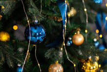 Fir Tree Decorated With Christmas Gold And Blue Balls. Close-up Of Beautiful Christmas Tree With Illuminated Garland And Blue Ribbons. Merry Xmas And Happy Holidays Greeting Card, New Year. Noel.