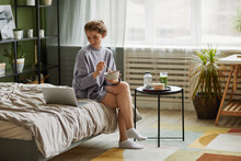 Young woman having breakfast and using laptop while sitting on bed in bedroom at home