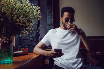 Wall Mural - African american man in a white t-shirt sits in a coffee shop and uses the phone. Mock-up.