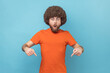© khosrork - Portrait of shocked surprised man with Afro hairstyle wearing orange T-shirt pointing down with fore fingers, looking at camera with open mouth. Indoor studio shot isolated on blue background.