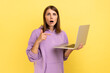 Portrait of amazed shocked young adult woman working on laptop compute, looking at camera and pointing at display, wearing purple hoodie. Indoor studio shot isolated on yellow background.