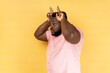 Side view of man wearing pink shirt showing bull horn gesture, looking at camera with hostile angry expression, bully male threatening to attack. Indoor studio shot isolated on yellow background.