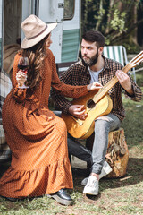 Wall Mural - Young beautiful loving couple travelling across country in the van. Millennial man and woman in a travel camper. Cozy atmosphere, vacations vibe. Drinking tea, playing guitar