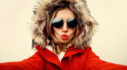 Canvas Print - Portrait of stylish woman blowing her lips sends air kiss stretching hand for taking selfie with smartphone wearing jacket with fur hood