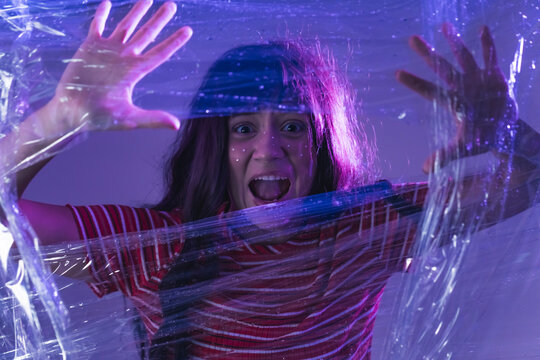 White teenage girl with dark hair and creative makeup trapped behind plastic wrap looking at camera screaming trying to escape. Artistic dramatic photography. Horizontal shot. High quality photo