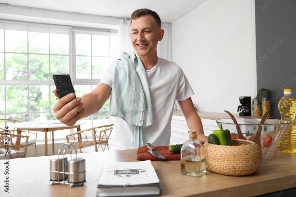 Young man with mobile phone taking selfie in kitchen