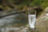 Fototapeta Łazienka - Fresh water pouring into glass on stone near stream. Space for text