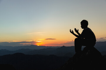 Wall Mural - Silhouette of a young man praying to God on the mountain at sunset background. Woman raising his hands in worship. Christian Religion concept.