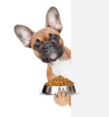 Poster - Funny french bulldog puppy holding bowl of dogs food behind empty white banner. isolated on white background
