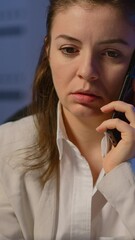 Poster - Vertical video: Tired freelancer woman having conversation on phone while working exhausted in business office late at night doing overtime. Focused employee using modern technology network wireless