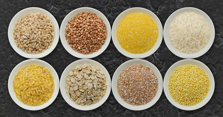 Different types of cereals for cooking: barley, buckwheat, corn, rice, bulgur, oatmeal, crushed wheat, millet in white bowls on dark background top view. Organic grains.  