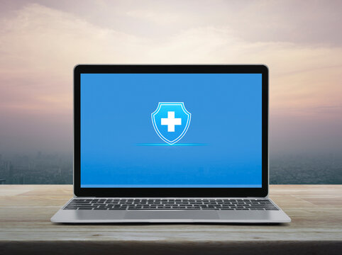 Cross shape with shield icon on modern laptop computer screen on wooden table over office city tower and skyscraper at sunset sky, vintage style, Business healthy and medical care insurance online con