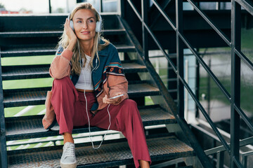 Wall Mural - Young attractive blonde woman listening to music on headphones while enjoying outside.