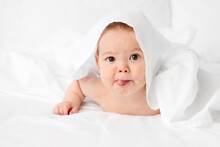 Funny Baby Lying On Bed Peeking Under White Sheet Sticking Tongue Out
