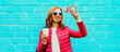 Portrait of happy smiling young woman taking selfie with smartphone wearing pink jacket on blue background