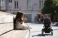 Disabled Woman With Reduced Mobility And Small Stature Sitting On A Wooden Bench, Consulting Social Networks On Her Cell Phone. Concept Handicap, Disability, Incapacity, Special Needs, Apps.