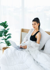 Wall Mural - Millennial Asian sexy female in casual comfort tanktop and shirt sitting under white thick warm blanket working with laptop computer drinking hot coffee after waking up in bedroom in morning at home