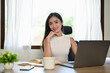 © bongkarn - Attractive millennial Asian businesswoman sits at her desk with her hand on her chin