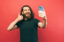 Photo Of Excited Young Bearded Hipster Man With Long Hair Taking A Selfie With Smartphone And Showing Thumb Up.