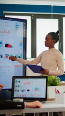 Wall Mural - Black woman worker explaning company statistics in front of colleagues, briefing group of employees. Multiethnical businesspeople working in professional startup financial company during conference