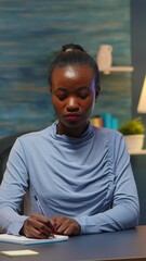 Poster - Vertical video: Black young business woman sitting at desk in living room workplace overtime and taking notes in notebook. African freelancer working from home late at night respecting deadline of