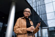 © Ivan Traimak - Business mature adult confident woman leader with a cup of coffee by a mobile phone against the background of the facade of an office skyscraper, business economic consultant concept