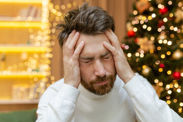 A man has a headache for Christmas, a close-up photo on New Year's Eve near the tree, a man holding his head with his hands, sick, sitting on the sofa at home alone.