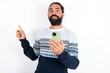 © Jihan - Smiling young bearded hispanic man wearing knitted sweater over white background pointing finger at blank space holding phone in one hand