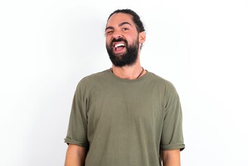 young bearded hispanic man wearing green t-shirt over white background winking looking at the camera
