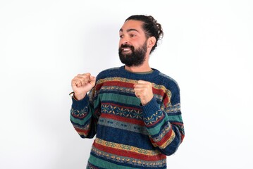 Caucasian man with beard wearing sweater over white background clenches fists and awaits for something nice happened looks away bites lips and waits announcement of results