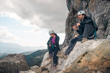 Concept: Adventure. Pair Of Climbers With Helmet And Harness. Resting Sitting On A Rock. Using The Smart Phone While Looking At The Horizon. Via Ferrata In The Mountains.