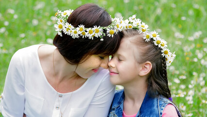 Summer, amidst a chamomile lawn, in the forest, a young woman, a brunette and a girl of seven, mother and daughter weave a wreath of chamomiles, laughing, trying on wreaths. High quality photo