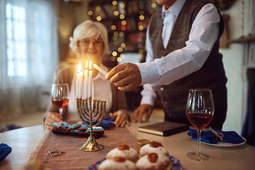 Wall Mural - Close up of mature man lighting menorah candles while celebrating Hanukkah with his wife at home.