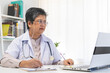 © KMPZZZ - Smile senior asian physician, general woman doctor wearing glasses and white uniform, mature female with stethoscope, working on laptop computer, sitting in medical office. Healthcare and medical.