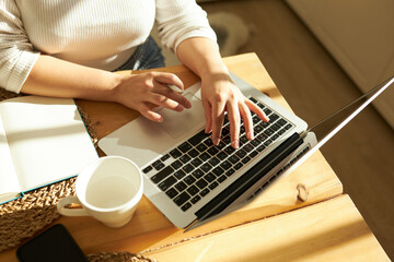 Cropped shot top view of female hands typing on keyboard of opened laptop at kitchen table with white coffee mug and notebook, studying online, checking e-mail, using learning application