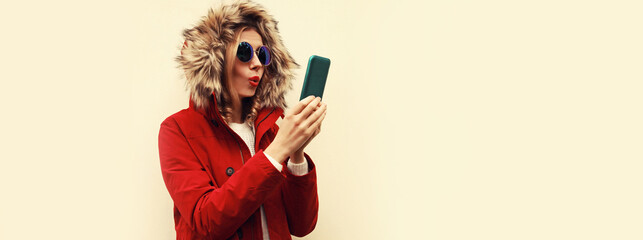 Wall Mural - Portrait of stylish young woman with smartphone wearing red jacket with fur hood, backpack on background, blank copy space for advertising text