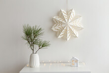 Christmas Cozy Winter Home Decor. New Year Interior Decorations. White Paper Snowflake On Wall, Green Pine Branch In Vase, Decorative Ceramic House, Glowing Garland Lights. Composition On The Dresser.