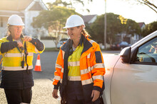 Two Smiling Women Road Workers Wearing White Helmet With Orange And Yellow Jacket On Sunset