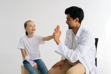 Wall Mural - Studio shot of cheerful little injured girl with broken hand wrapped in plaster bandage giving high five to friendly male doctor at checkup meeting, on white isolated background.