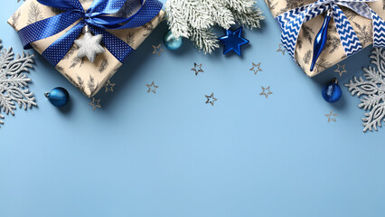 Christmas frame top border made of fir tree branches, gift boxes, silver blue stars, balls over blue background. Flat lay, top view. Xmas banner mockup
