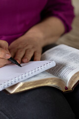 Wall Mural - Senior Christian woman writing in a notebook with an open Holy Bible Book in her lap. Vertical shot, a close-up. Selective focus. Writing biblical verses, reading and studying Scriptures concept.